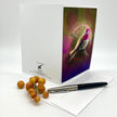 A art print greeting card featuring a hummingbird perched on branches with a glowing circular motif and pink-toned background, styled on a white paper envelope and surface with a white envelope and a black pen nearby.