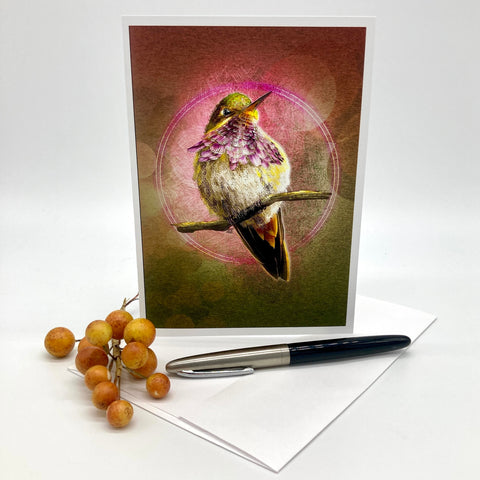 An art greeting card featuring a detailed illustration of a hummingbird perched on a branch with purple flowers, displayed on a white surface next to a black and silver pen and a small cluster of brown berries.