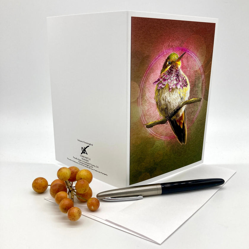 A white greeting card set displayed on a light gray background, featuring a front cover illustration of a hummingbird with purple flowers. A matching white envelope and a black-and-silver pen are placed in front, along with a small cluster of orange-brown grapes.