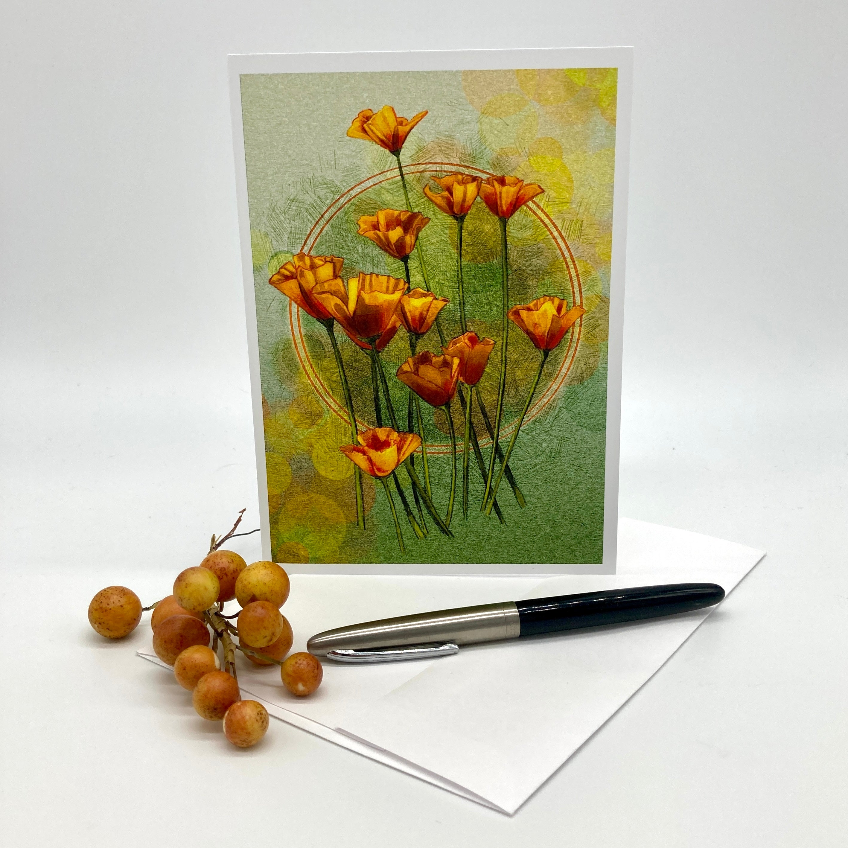 A vertical greeting card featuring orange flowers on a textured green and yellow background, displayed upright with an envelope and a black-and-silver pen on a white surface. A small cluster of round tan berries sits beside the card.