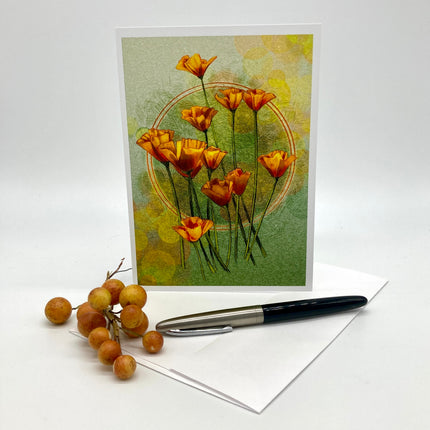 A vertical greeting card featuring orange flowers on a textured green and yellow background, displayed upright with an envelope and a black-and-silver pen on a white surface. A small cluster of round tan berries sits beside the card.