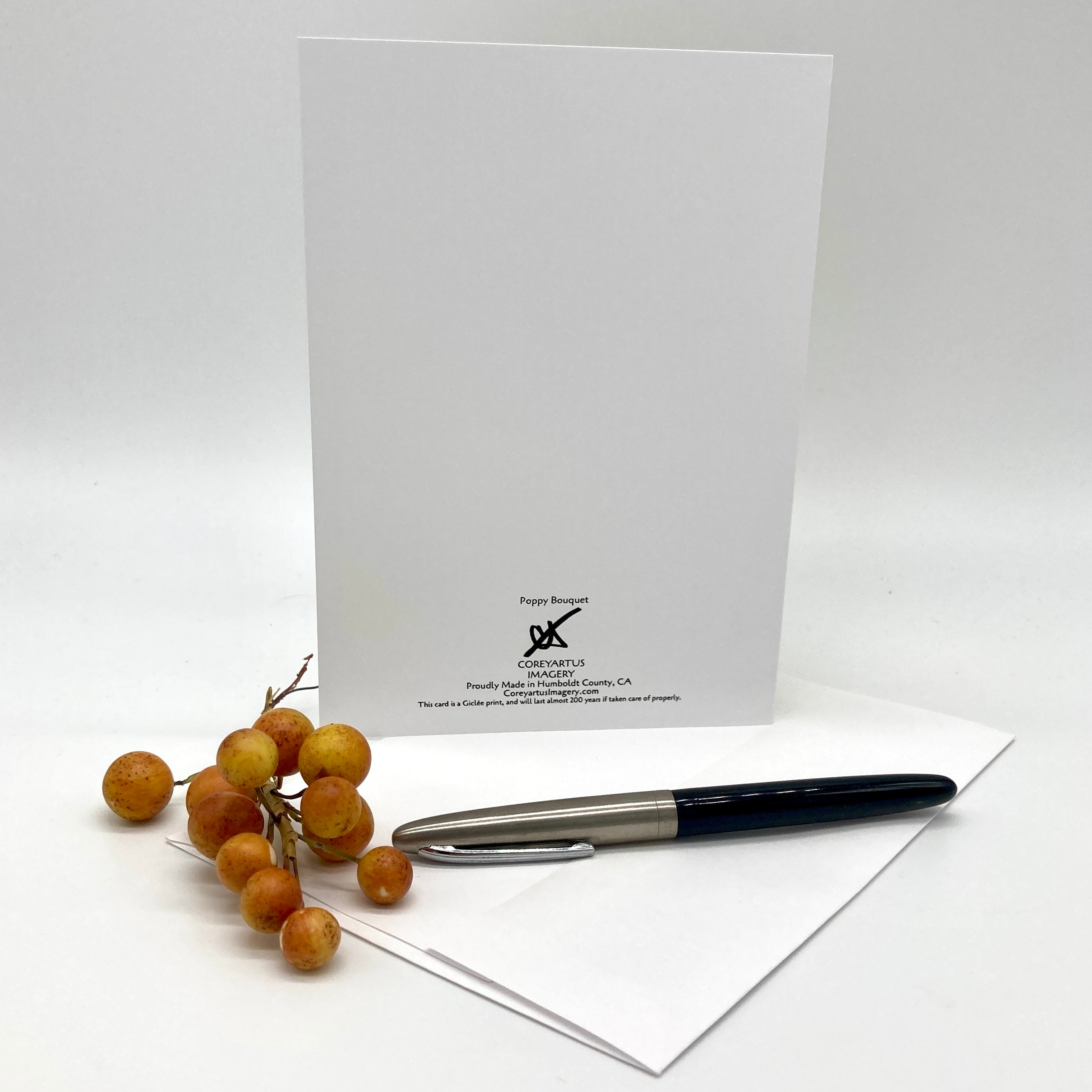 A white greeting card with black printed text and logo stands upright on a white surface, with an envelope and a black-and-silver pen in front. A small cluster of tan berries sits to the left.