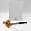 A white greeting card with black printed text and logo stands upright on a white surface, with an envelope and a black-and-silver pen in front. A small cluster of tan berries sits to the left.