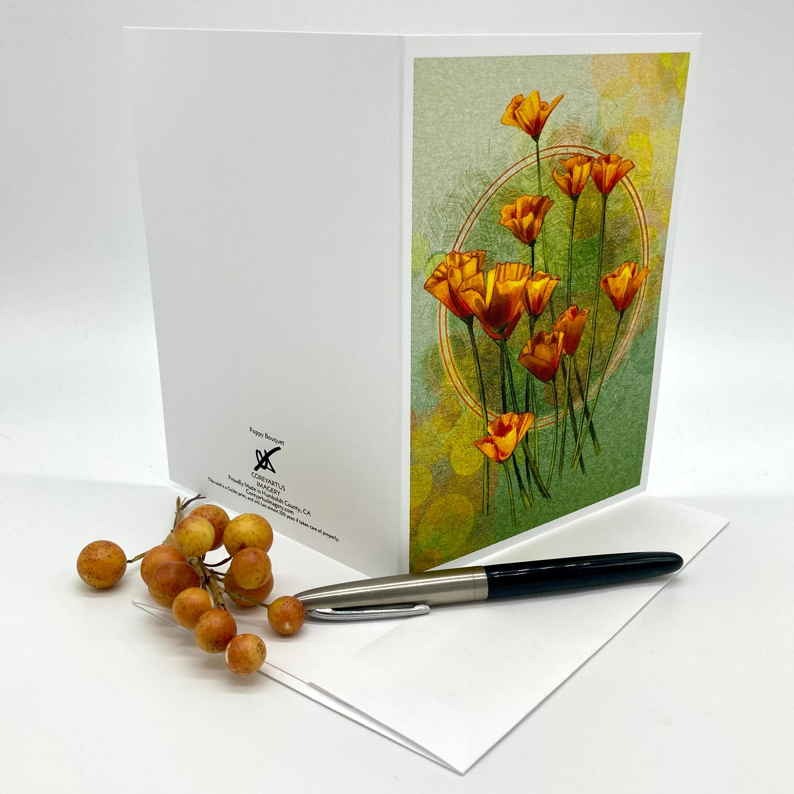 A white greeting card with a floral illustration of orange flowers on a green and yellow textured background, displayed upright with an envelope and a black pen on a white studio background. Small orange-brown berries are placed beside the card.