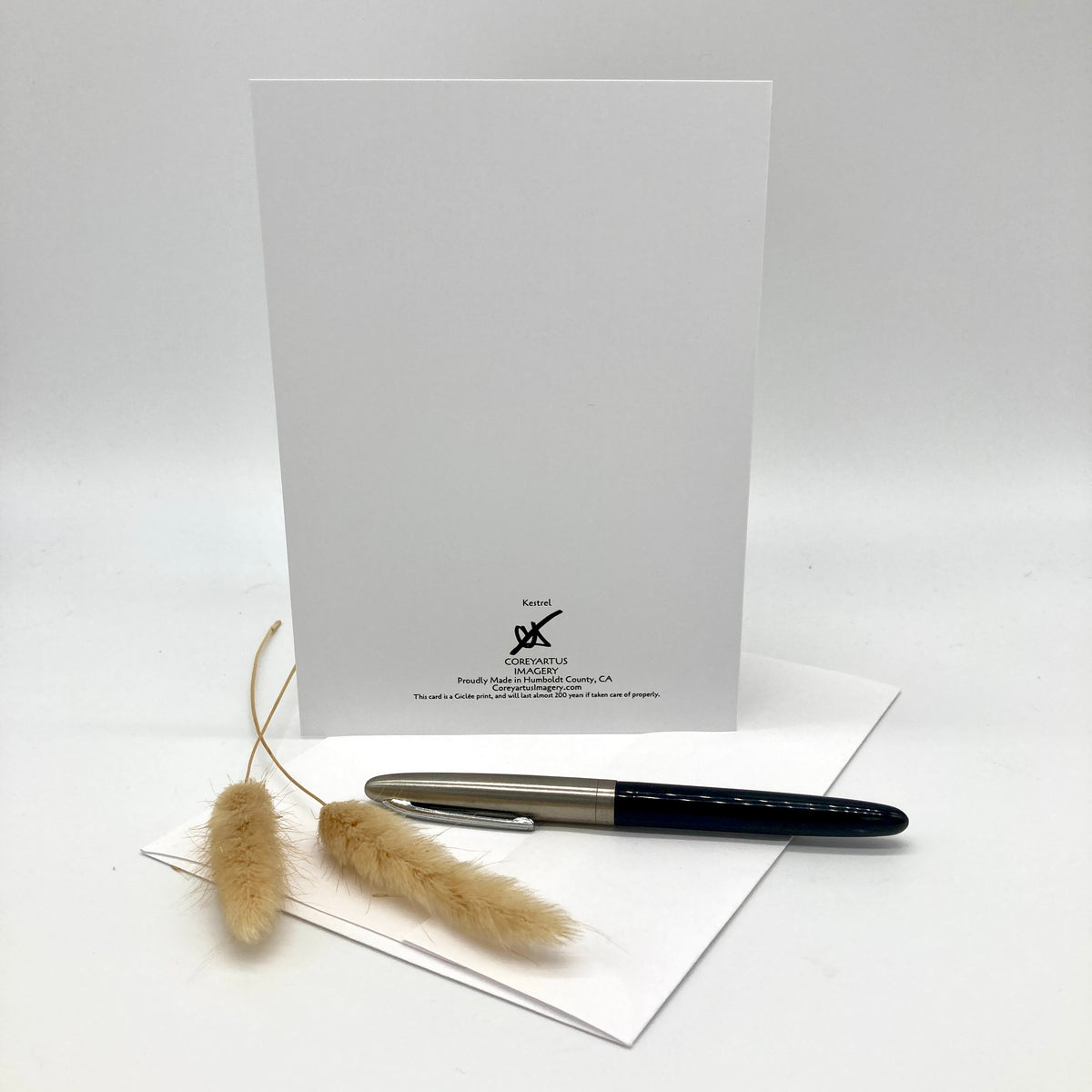 A white greeting card with a black printed logo and text stands upright on a white surface, with a black and silver pen resting on a folded white envelope in front and dried bunny tail grass beside it.
