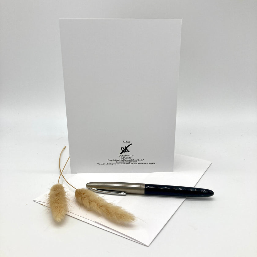 A white greeting card with a black printed logo and text stands upright on a white surface, with a black and silver pen resting on a folded white envelope in front and dried bunny tail grass beside it.