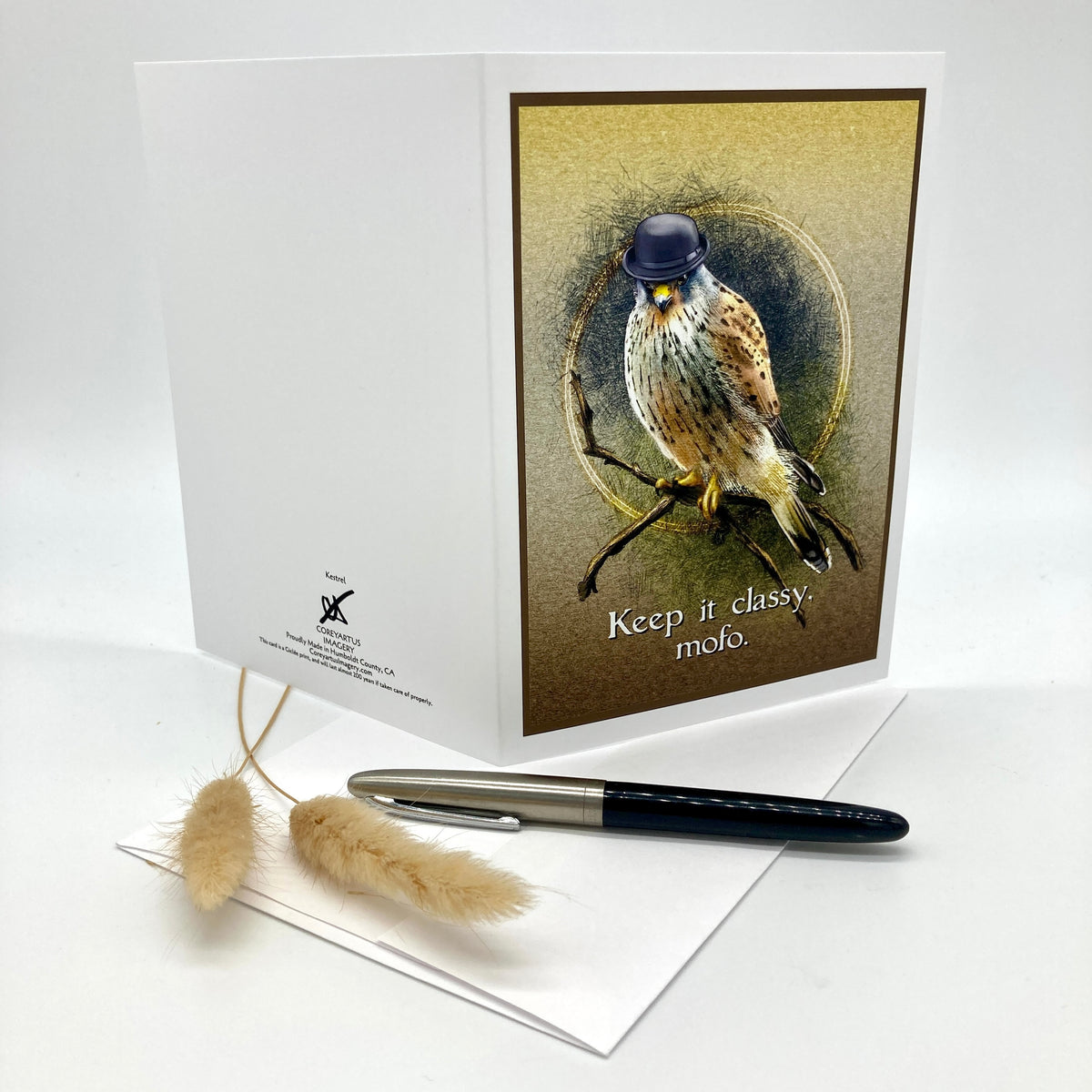 A white greeting card with a printed illustration of a bird wearing a small black hat, perched on a branch with the text 'Keep it classy, mofo.' The card is shown standing upright on a white surface, with a black and silver pen resting on top and dried decorative grasses in front.