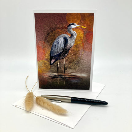 A vertical art print or greeting card featuring a painted heron standing in shallow water, displayed upright on a white surface with a white envelope, a black pen, and two dried beige grasses in front.