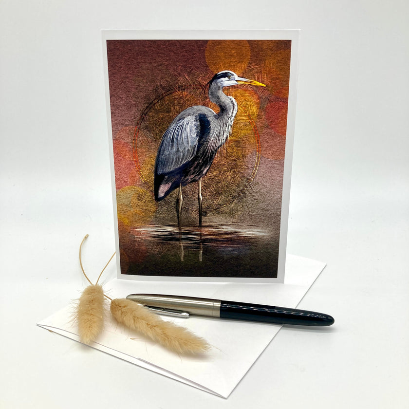 A vertical art print or greeting card featuring a painted heron standing in shallow water, displayed upright on a white surface with a white envelope, a black pen, and two dried beige grasses in front.