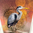 A close-up of a textured art print or painting depicting a gray heron standing in shallow water, with a warm abstract background in rust, orange, and brown tones.