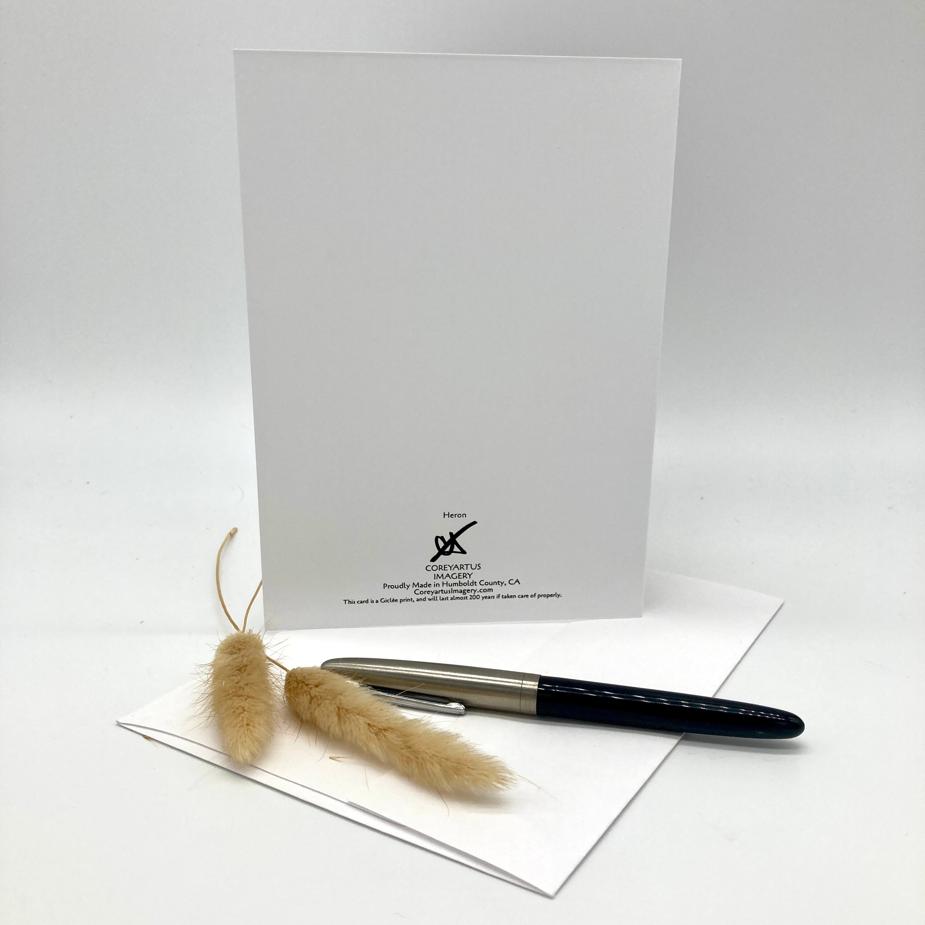 A white stationery card with black printed text and logo stands upright on a white surface, with a black and silver pen resting on a white envelope in front and two dried beige grasses as styling props. The card shows the brand name 'The Art of Writing' and 'Pensiero' near the bottom.