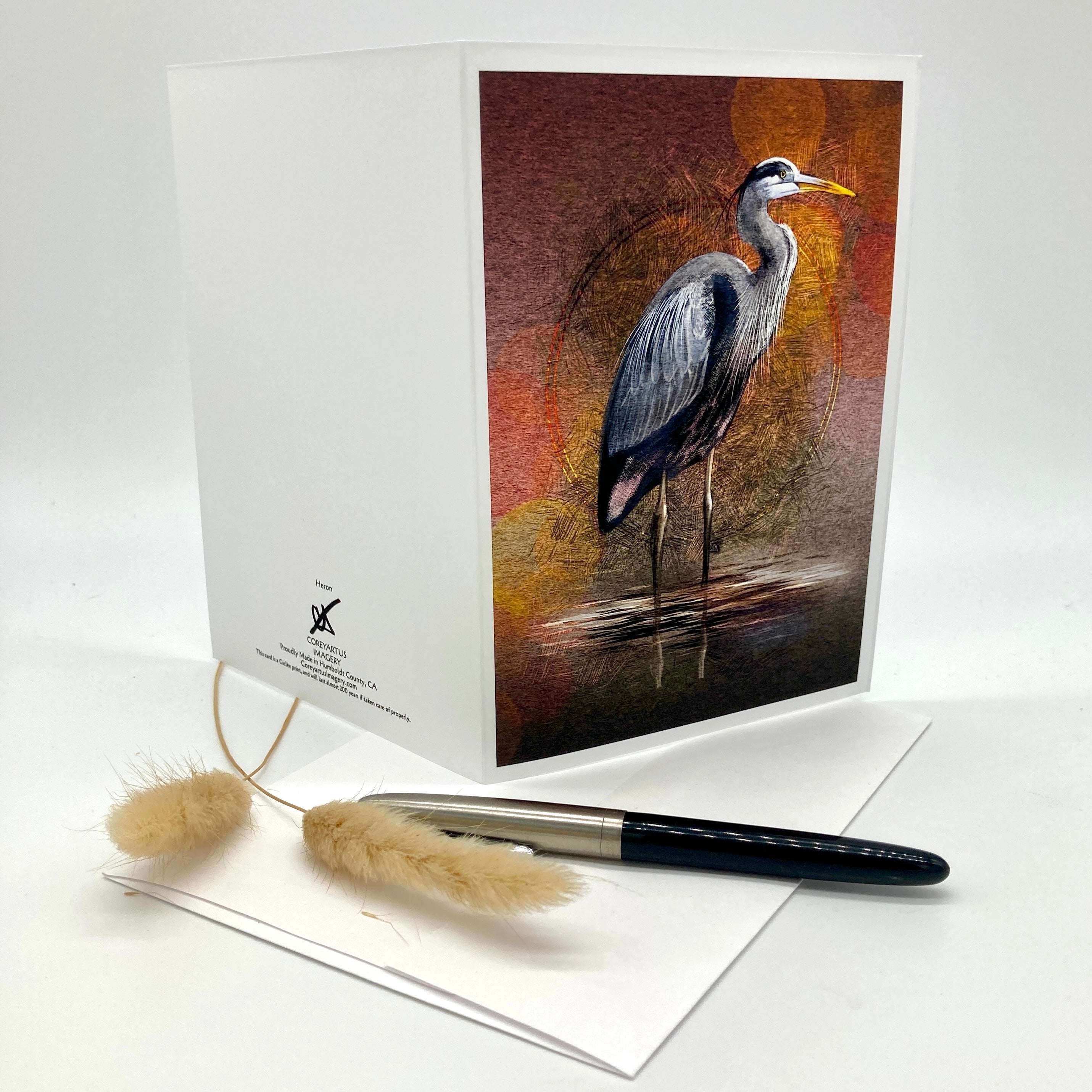 A white greeting card with a printed illustration of a heron on the front, displayed upright with a matching white envelope and a black pen on a clean white background.