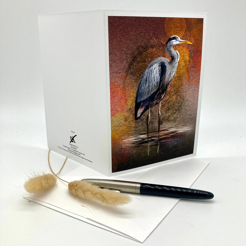 A white greeting card with a printed illustration of a heron on the front, displayed upright with a matching white envelope and a black pen on a clean white background.