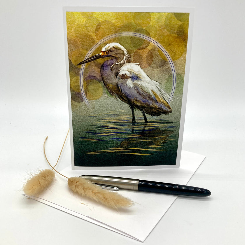 A vertical art print or postcard featuring a white egret standing in reflective water, with a yellow-green abstract background and a thin circular frame around the bird. The print is displayed upright on a white surface with a white envelope, a black pen, and two dried bunny tails as styled props.