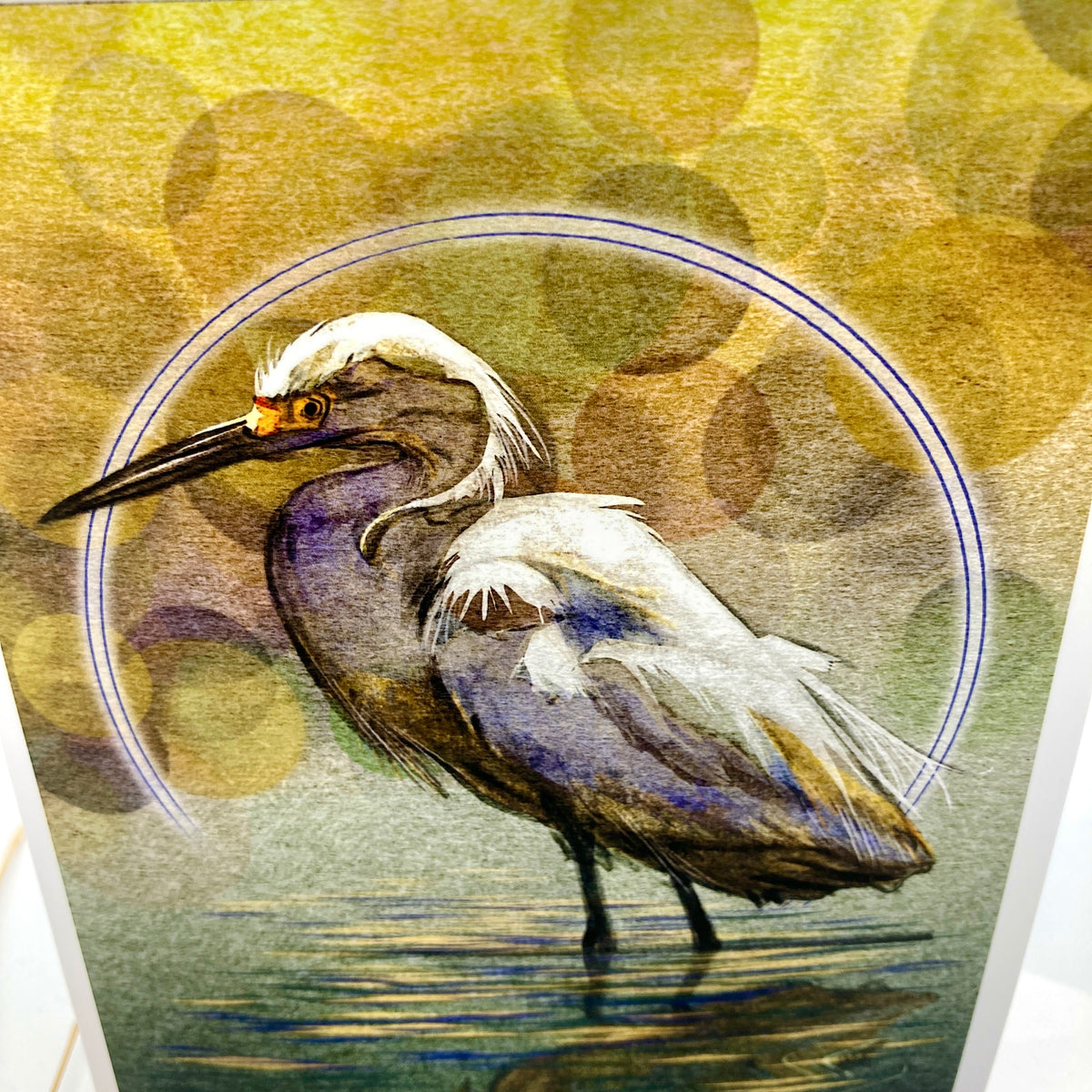 A close-up of a printed artwork showing a heron standing in shallow water, with a circular blue outline behind it and a warm yellow-green bokeh-style background. The print is photographed at an angle, with part of the white page border visible on the right.