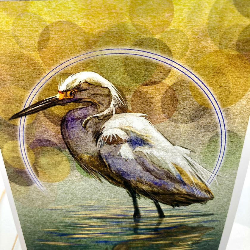 A close-up of a printed artwork showing a heron standing in shallow water, with a circular blue outline behind it and a warm yellow-green bokeh-style background. The print is photographed at an angle, with part of the white page border visible on the right.