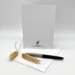 A white stationery card with a black printed logo and address stands upright on a white surface, with a black and silver pen resting across the bottom and two dried beige grasses in the foreground on a light gray studio background.