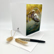 A white greeting card standing upright on a white surface, featuring a printed illustration of a white heron standing in water against a yellow-green background. A black pen and two dried beige grass stems are placed in front of the card.