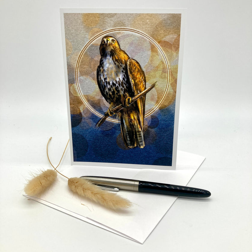 A vertical greeting card featuring a detailed illustration of a hawk perched on a branch against a blue and gold background, displayed on a white envelope with a black pen and two dried bunny tails on a white studio background.