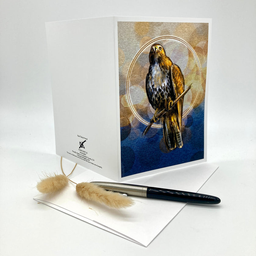 A white greeting card featuring an illustration of a hawk in a circular frame against a blue and beige background, displayed with a black and silver pen and two dried grass stems on a white surface.