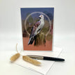 An art print or greeting card featuring a detailed bird perched on a tree stump, displayed upright on a white envelope with a black pen and two dried grass stems in front, against a clean white background.
