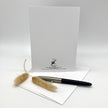 A white greeting card with a black printed logo and address stands upright on a white surface, with a black and silver pen resting on an envelope in front and two dried beige grasses as styling props.