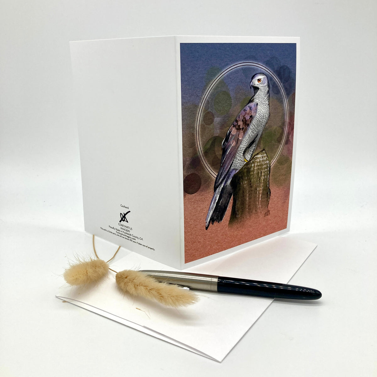 A white greeting card standing upright on a white surface, featuring an illustrated bird with blue, gray, and brown tones inside an oval frame on the front. A black pen and two beige dried grass stems rest in front of the card.