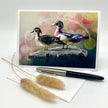 A greeting card featuring two painted ducks on a rock, displayed upright on a white surface with a pen and dried grass stems in front.