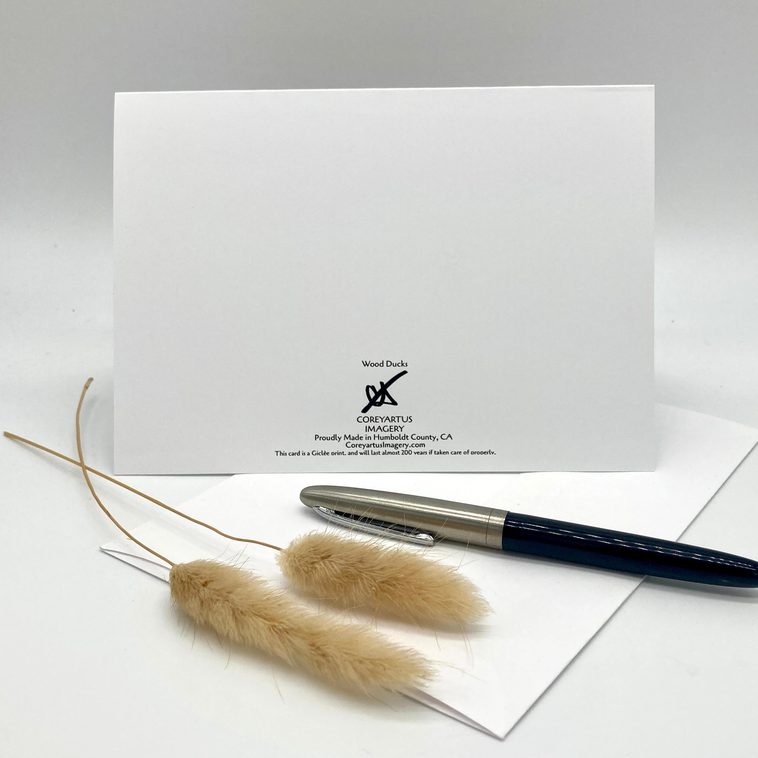 A white stationery card or envelope with the Wood Duke Corporation logo printed on it, styled with a dark blue and silver fountain pen and two dried pampas grass stems on a light gray background.