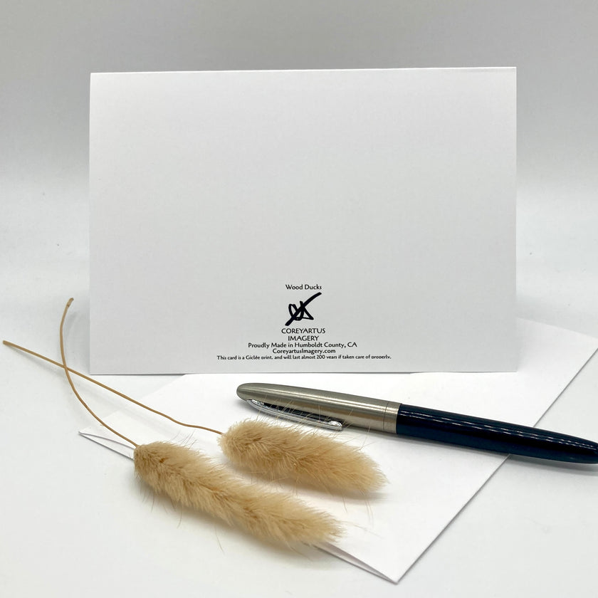 A white stationery card or envelope with the Wood Duke Corporation logo printed on it, styled with a dark blue and silver fountain pen and two dried pampas grass stems on a light gray background.