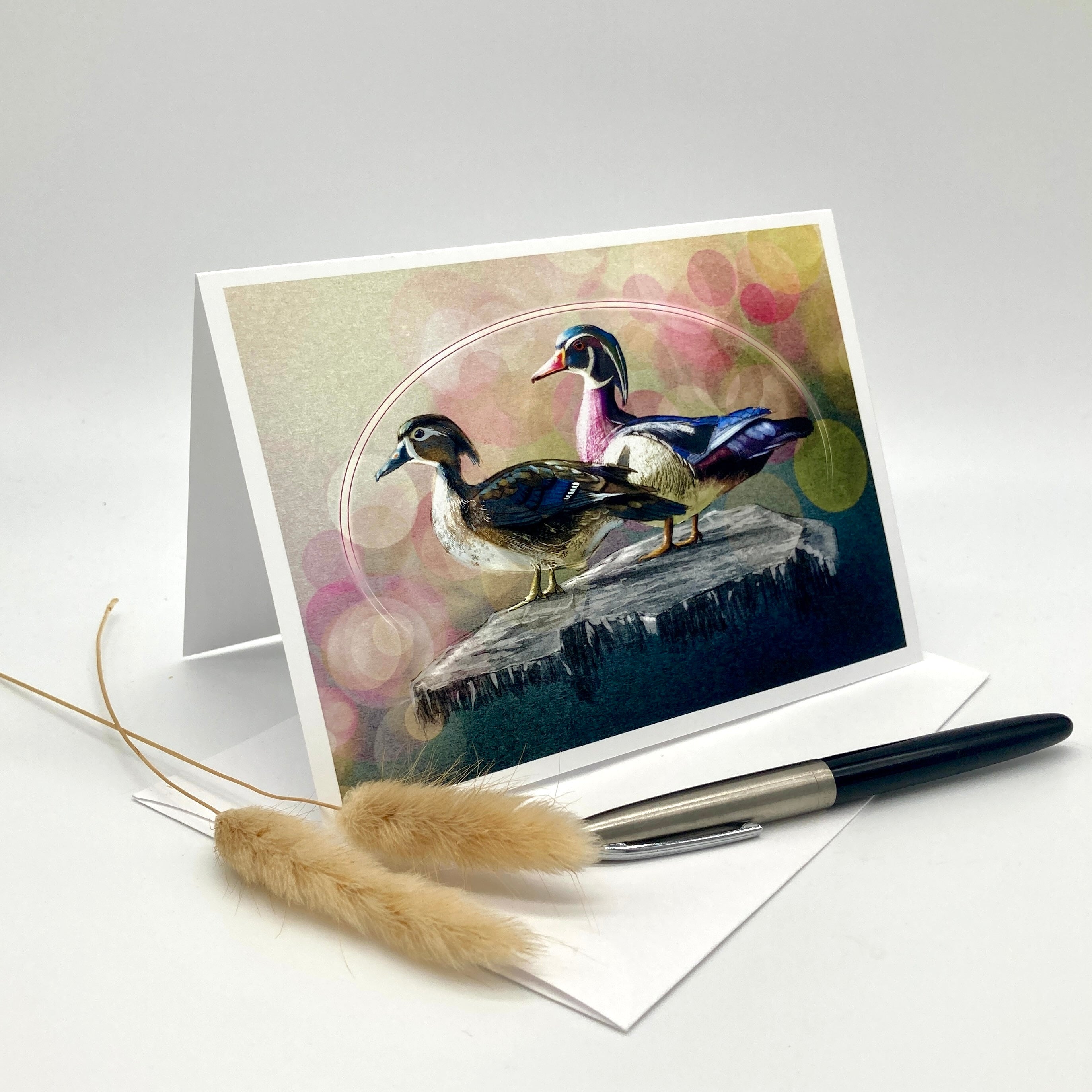 A greeting card with a colorful illustration of two ducks standing on a rock, displayed in a white stand. A white envelope, a black pen, and two dried pampas grass stems are arranged on a light gray studio background.
