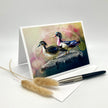 A greeting card with a colorful illustration of two ducks standing on a rock, displayed in a white stand. A white envelope, a black pen, and two dried pampas grass stems are arranged on a light gray studio background.