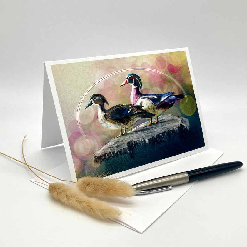 A greeting card with a colorful illustration of two ducks standing on a rock, displayed in a white stand. A white envelope, a black pen, and two dried pampas grass stems are arranged on a light gray studio background.
