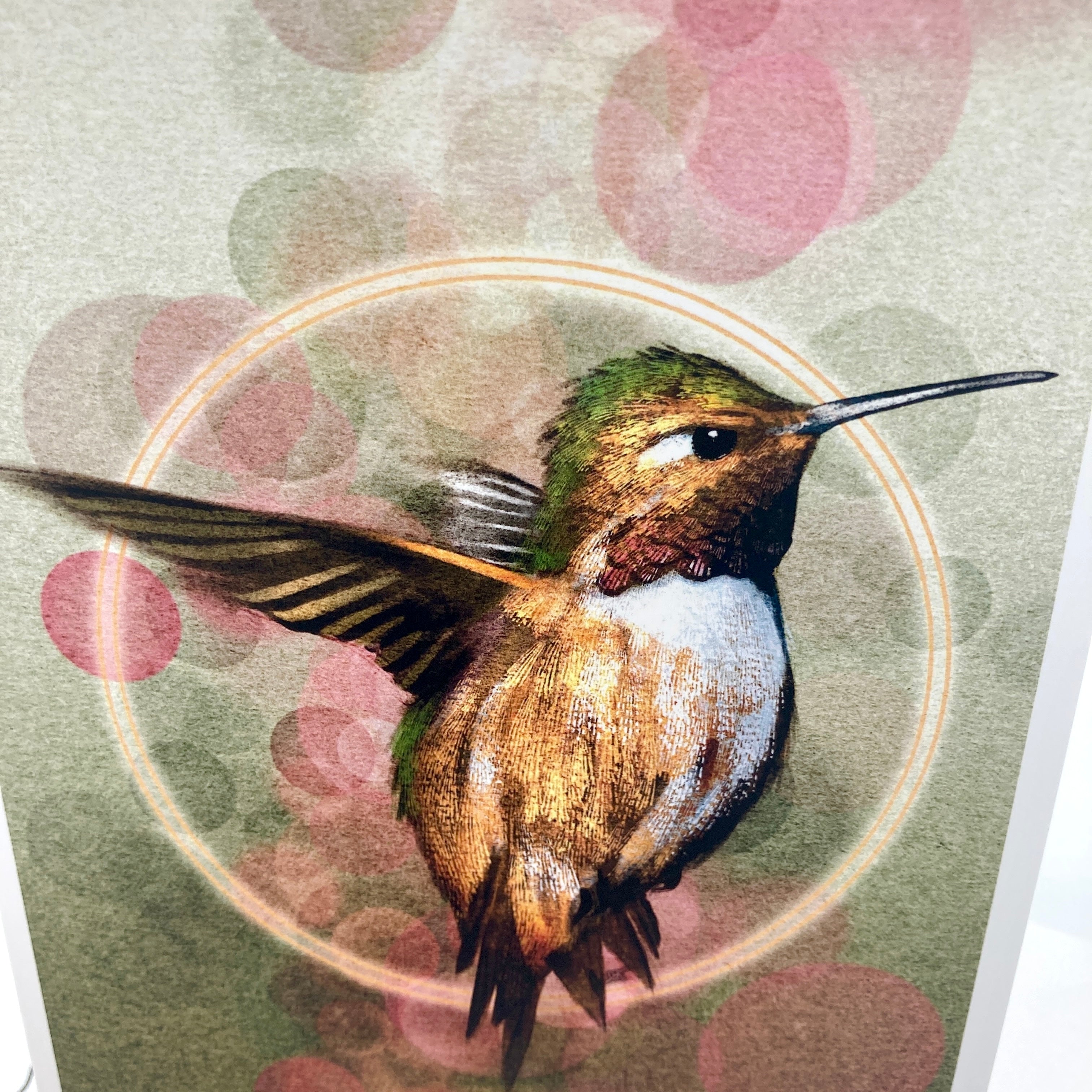 A close-up of a printed artwork featuring a hummingbird in motion over a soft-focus background with pink and green bokeh circles.