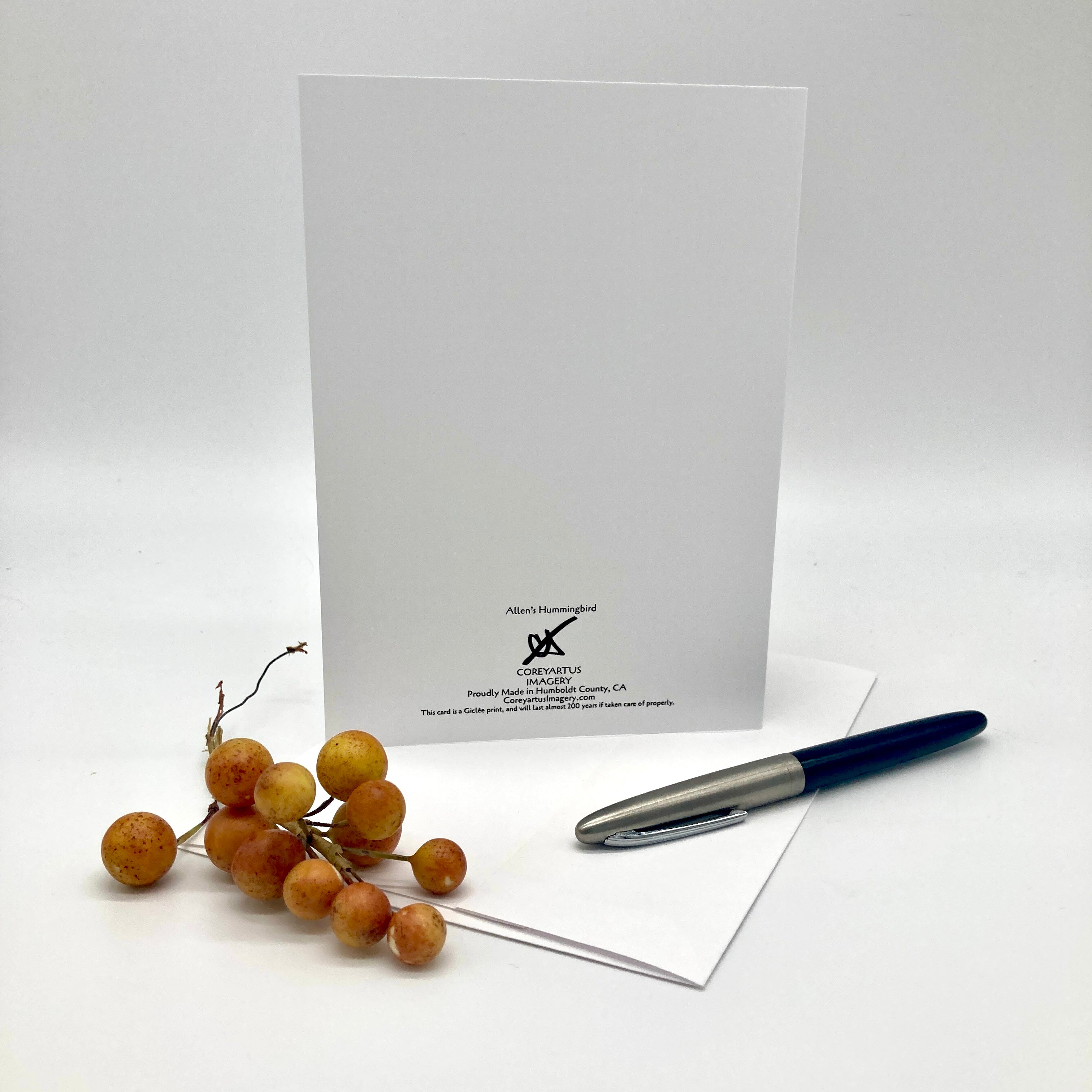 A white greeting card with black printed text and logo stands upright on a white surface, with a silver and navy pen resting on an envelope in front and a small cluster of orange-brown berries to the left. The card is branded Allen's Organized.
