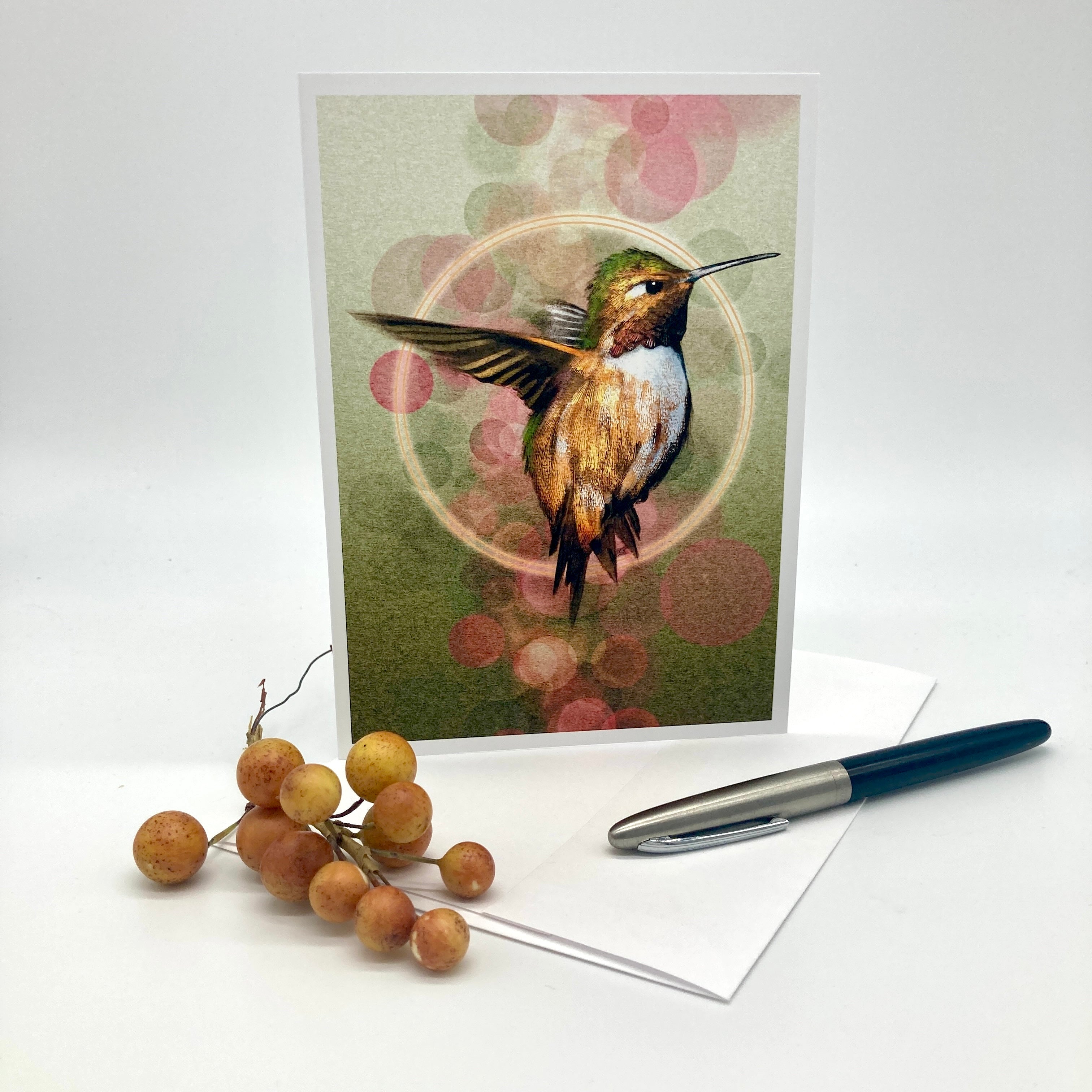 A greeting card featuring a detailed illustration of a hummingbird, displayed upright on a white surface with a white envelope, a blue and silver pen, and a small decorative cluster of orange berries.