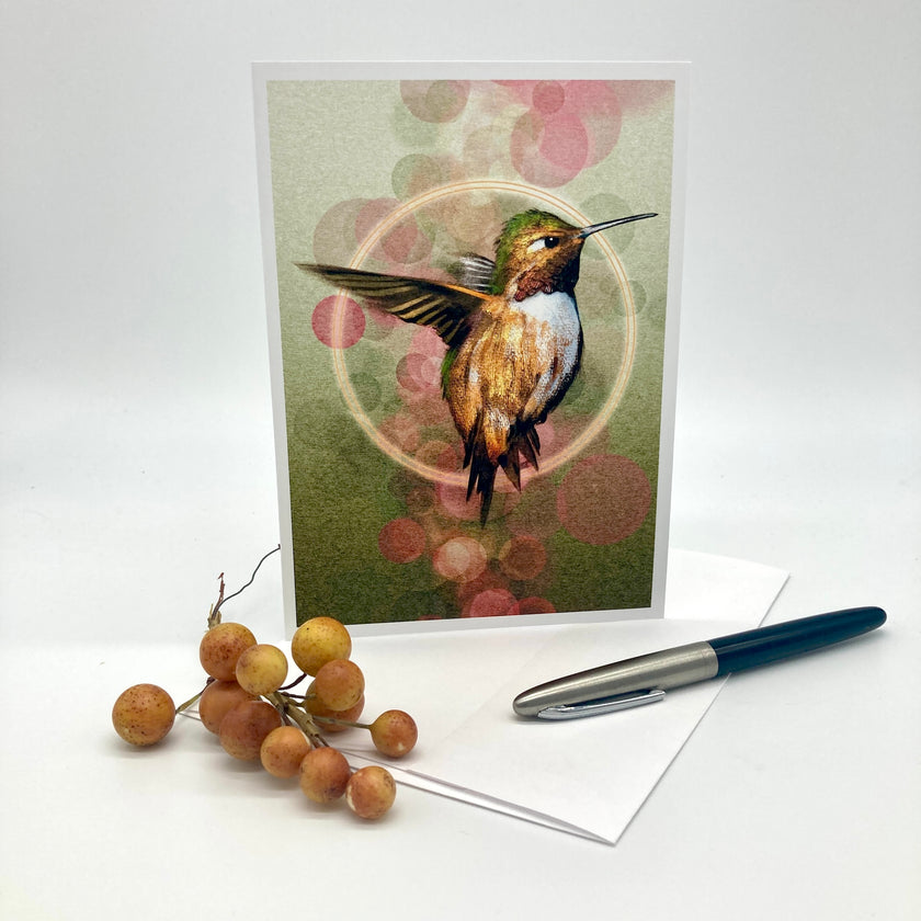 A greeting card featuring a detailed illustration of a hummingbird, displayed upright on a white surface with a white envelope, a blue and silver pen, and a small decorative cluster of orange berries.