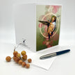 A white greeting card featuring an illustration of a hummingbird with green, gold, and pink tones, displayed on a white background with a pen and small orange berries as props.