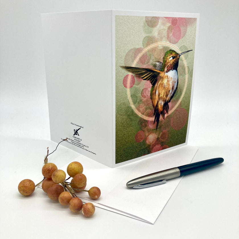 A white greeting card featuring an illustration of a hummingbird with green, gold, and pink tones, displayed on a white background with a pen and small orange berries as props.