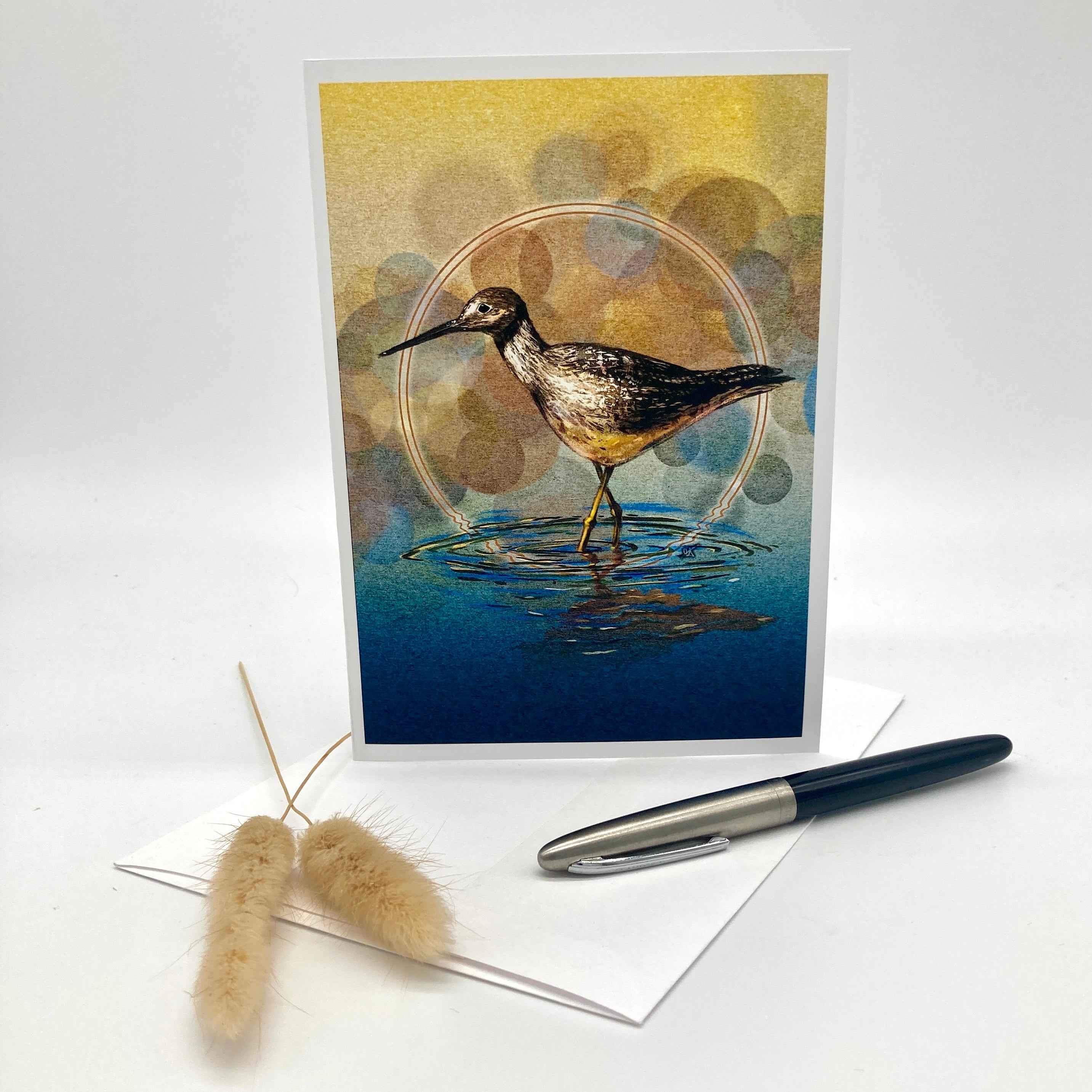 A vertical art print or greeting card featuring a bird standing in shallow water, styled on a white surface with a white envelope, a black and silver pen, and dried bunny tail grass. The artwork has a yellow and blue background with bokeh-style circles.