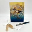 A vertical art print or greeting card featuring a bird standing in shallow water, styled on a white surface with a white envelope, a black and silver pen, and dried bunny tail grass. The artwork has a yellow and blue background with bokeh-style circles.
