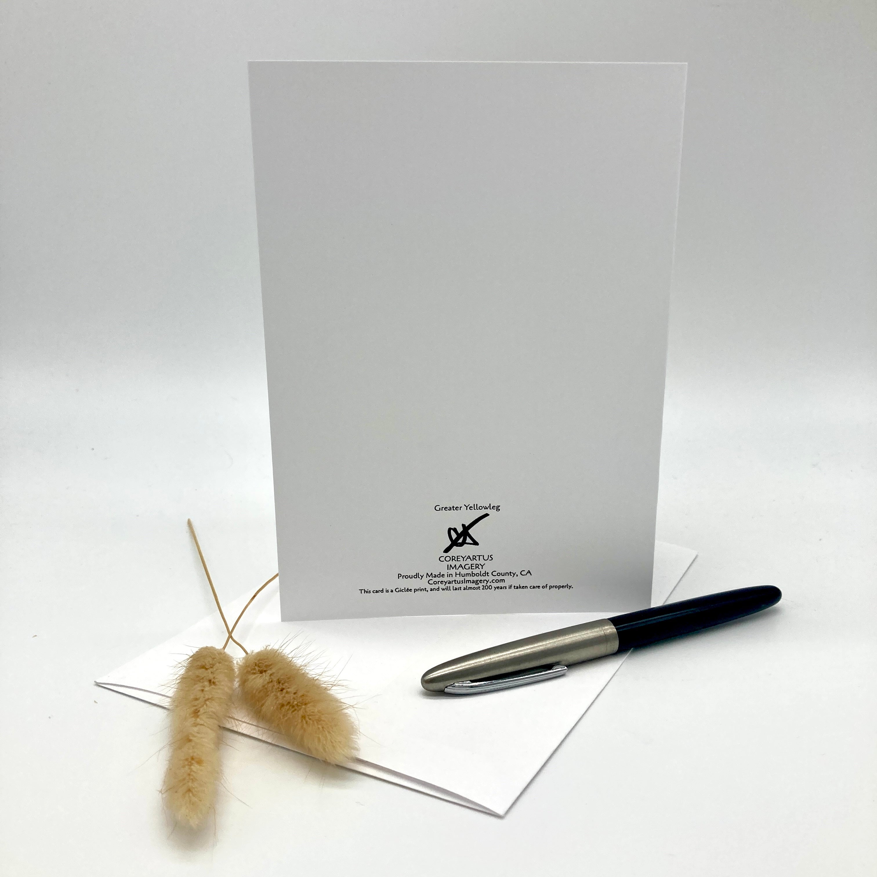 A white greeting card with black printed text and logo stands upright on a white surface, with a silver and black pen and two dried beige grass stems placed in front on a white background.