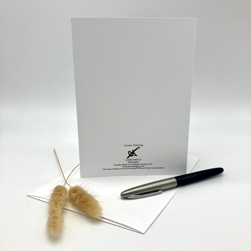 A white greeting card with black printed text and logo stands upright on a white surface, with a silver and black pen and two dried beige grass stems placed in front on a white background.
