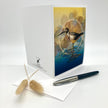 A white greeting card with a printed illustration of a wading bird in blue and gold tones, displayed upright on a white surface with a matching envelope, a black and silver pen, and two dried beige grasses.