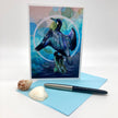 A square greeting card featuring a colorful illustration of a cormorant bird with wings spread, standing on a rock in front of blue water. The card is displayed upright on a light blue envelope with two seashells and a black-and-silver pen on a white studio background.
