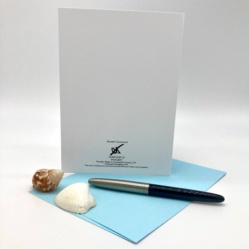 A white greeting card standing upright on a light blue envelope, styled with two seashells and a dark blue pen on a clean white background. The card features black printed text and logo for 'Breathe Beautifully'.