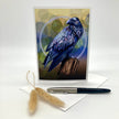A vertical greeting card featuring a detailed illustration of a black bird perched on a branch against a yellow, blue, and brown textured background, displayed with a white envelope and a black-and-silver pen on a white studio background.