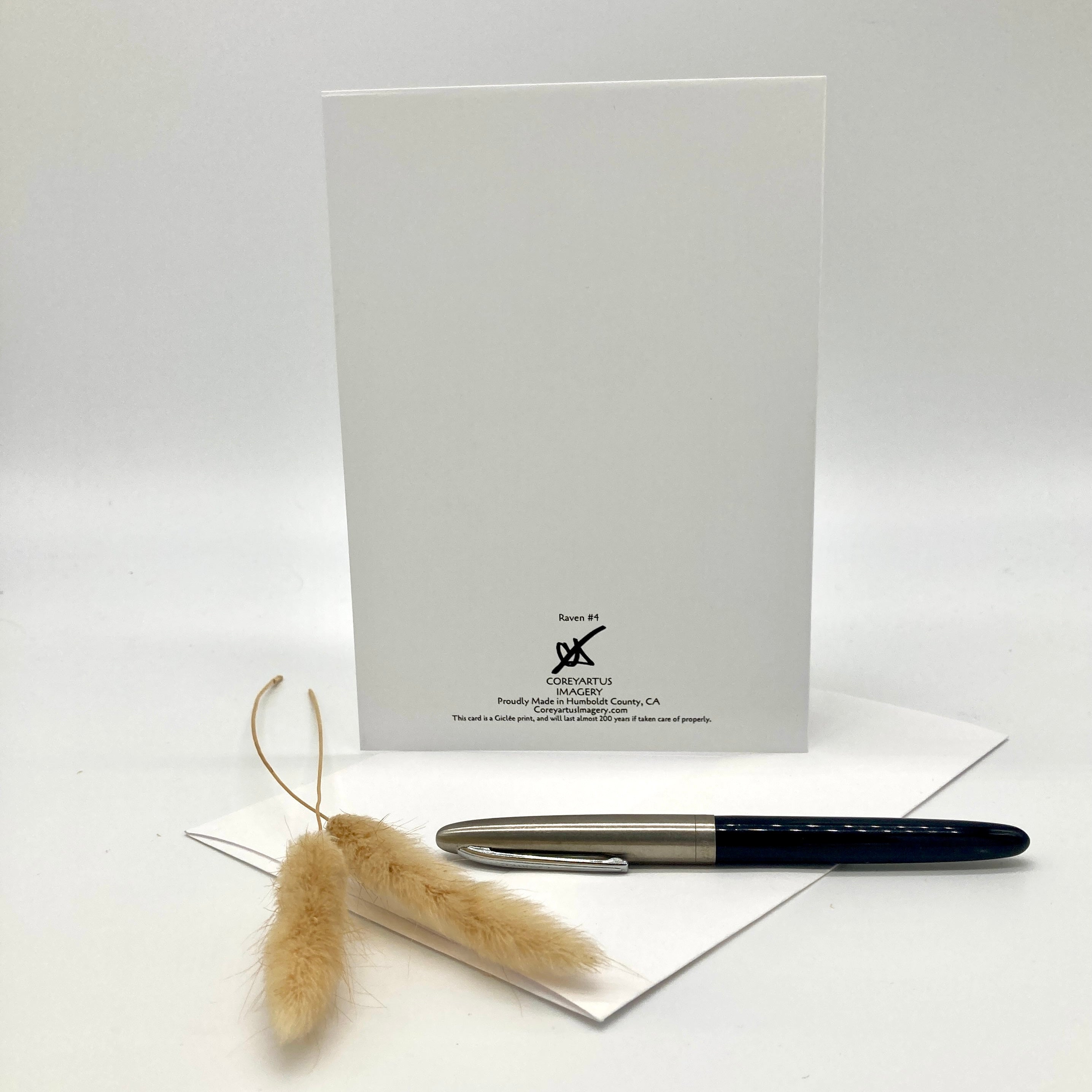 A white card or envelope standing upright on a white envelope, with a black and silver pen resting on the blue paper and two dried bunny tail grass stems in front. Small printed branding is visible near the bottom of the white card.