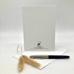 A white card or envelope standing upright on a white envelope, with a black and silver pen resting on the blue paper and two dried bunny tail grass stems in front. Small printed branding is visible near the bottom of the white card.