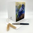 A white greeting card with a detailed illustration of a blue bravenon the front, displayed alongside a light blue envelope, a black and silver pen, and two dried bunny tail grass stems on a white background.